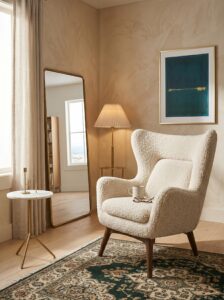 Wooden frame cream armchair with rounded cushions, beige curtains, botanical wall art, woven rug, ceramic decor, and side table