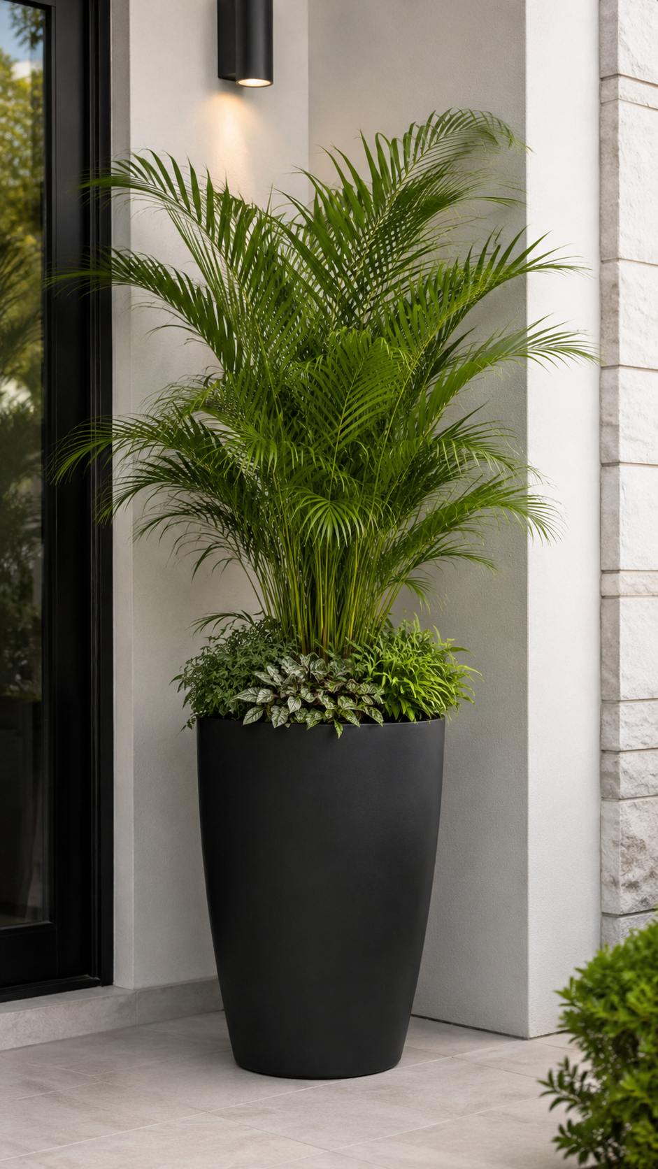Fresh front door planter with large matte black pot, palm-style greenery, mixed base plants, pale gray wall, stone detail, tiled floor, and black door